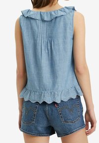 Kvinna i ärmlös ljusblå blus med volanger och broderad nederkant, samt jeansshorts med stickade bakfickor, sedd bakifrån.