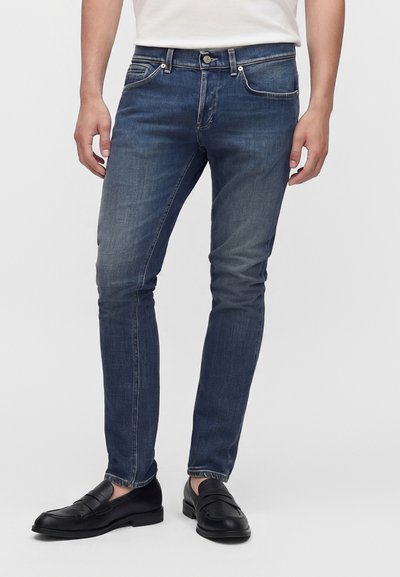 Homme portant un jean slim bleu foncé légèrement délavé et des mocassins en cuir noir, debout devant un fond clair uni.