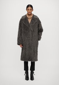 Tamsiai pilkas dirbtinės kailio paltas su tekstūruota paviršiumi, atvira priekyje ir plačiais apykaklėmis. Apsivilktas virš smėlio spalvos megztinio su aukštu kaklu ir juodomis kelnėmis.