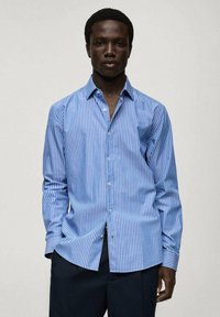 Camicia a righe blu e bianche con maniche lunghe, colletto classico e una texture del tessuto liscia. Abbinata a pantaloni scuri.