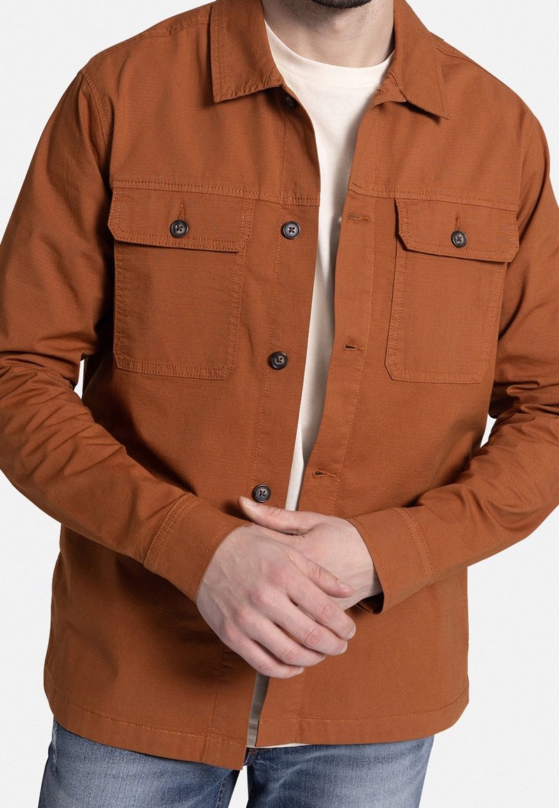 Homme portant une veste-chemise couleur rouille par-dessus un t-shirt blanc et un jean bleu, les mains jointes devant.