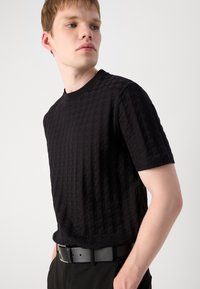 Fekete textúrájú, rövid ujjú póló, rácsos mintával, kerek nyakkivágással, és hozzá illő fekete övvel, fekete nadrággal viselve, világos háttér előtt.