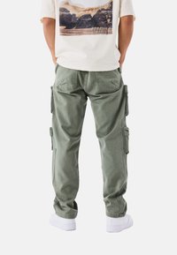 Pantalones cargo verdes de algodón con múltiples bolsillos laterales, ajuste relajado y puños en los bajos. Combinados con una camiseta color crema y zapatillas blancas.