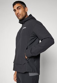 Chaqueta impermeable negra con capucha, con cierre de cremallera y bolsillos laterales. El material es elegante con un acabado mate y un logotipo en contraste.