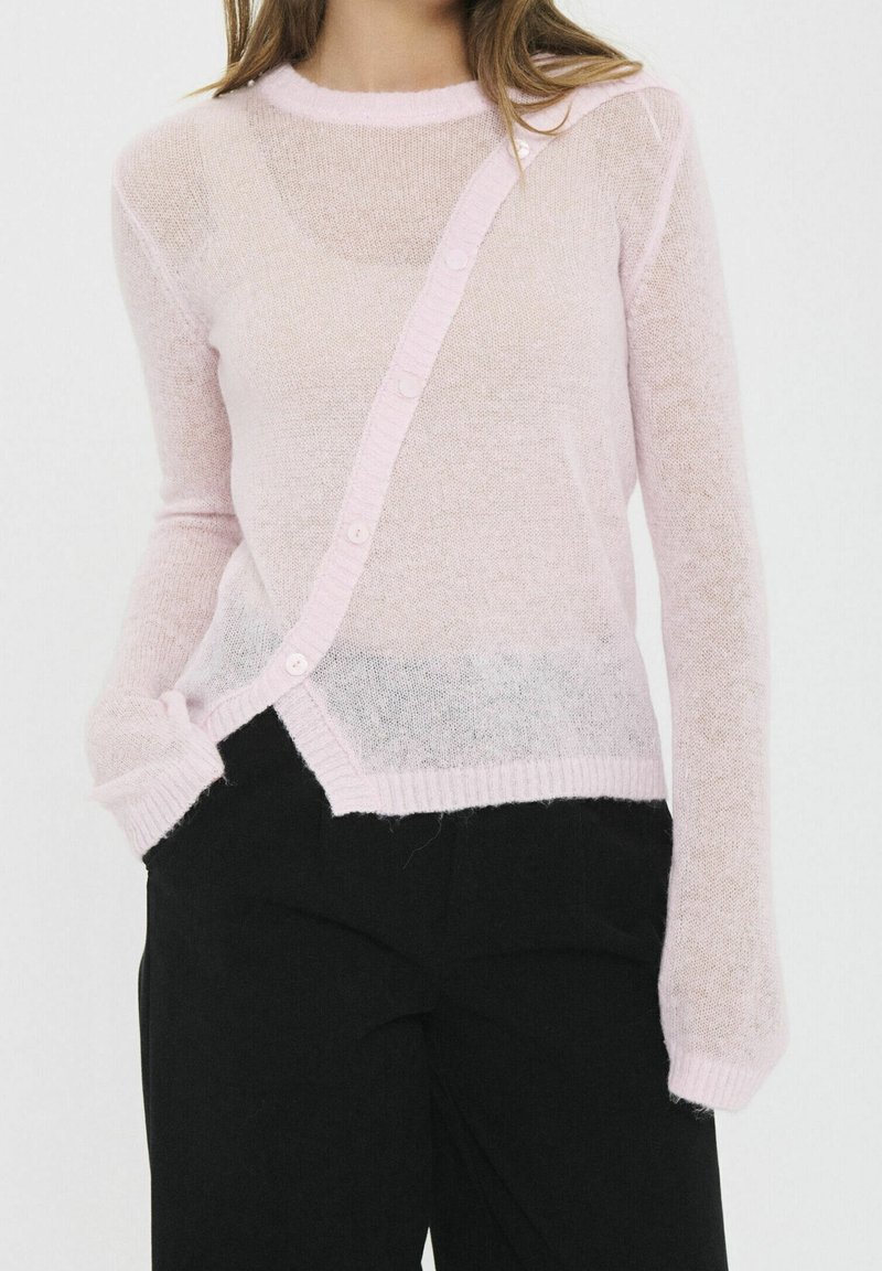 Femme portant un pull en maille léger rose clair et transparent avec des boutons en diagonale sur le devant, associé à un pantalon noir. Une main dans la poche.