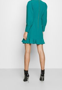 Robe bleu sarcelle avec un ourlet à volants, des manches bouffantes et une fermeture éclair centrale à l'arrière ; associée à des bottines noires en cuir verni avec des talons transparents.