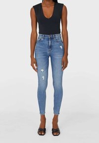 Musta bodysuit syvällä V-kaula-aukolla, yhdistettynä vaaleansinisiin korkea vyötäröisiin skinny-farkkuihin, joissa on kulutettuja yksityiskohtia ja revityt helmät. Mustat sandaalit.