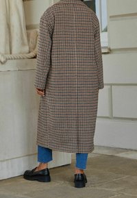 Lange, bruine geruite jas met een rechte snit, voorzien van grote ruiten en een kraag. Draagt over blauw cropped jeans en zwarte loafers met een dikke zool.