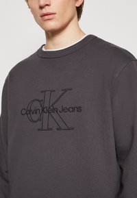 Grijs sweatshirt met een ronde halslijn, voorzien van zwarte geborduurde tekst "Calvin Klein Jeans" en een prominent "CK" logo aan de voorkant.