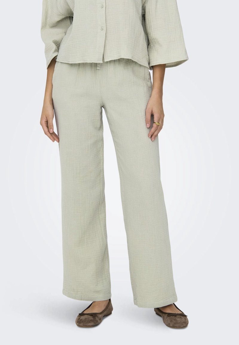Femme portant un pantalon large beige clair texturé, une chemise boutonnée assortie et des ballerines marron, debout les mains détendues le long du corps.