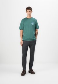 Grøn bomulds t-shirt med korte ærmer og et blåt grafisk tryk på brystet, parret med mørkegrå slim-fit bukser og brune snørebåndssko.