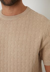 Beige gestrickter Pullover mit kurzen Ärmeln, der ein strukturiertes vertikales Streifenmuster und einen gerippten Rundhalsausschnitt aufweist, aus weichem Material gefertigt.