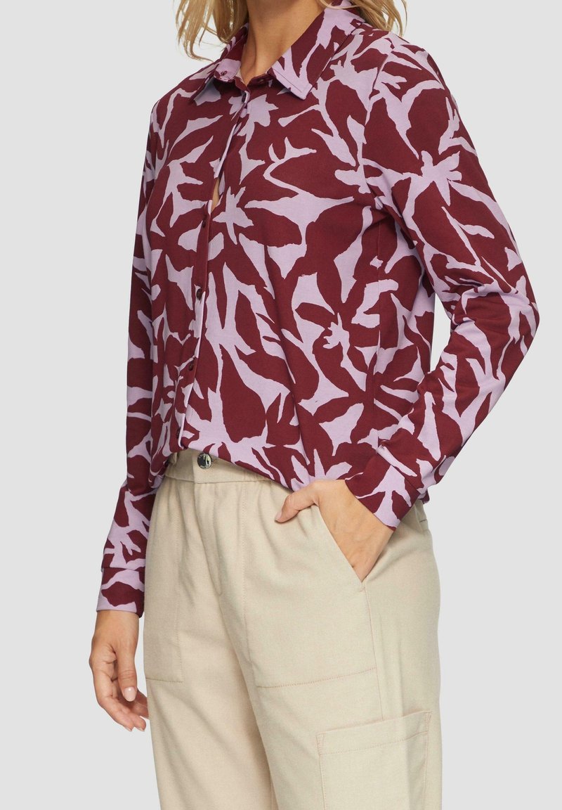 Femme portant une chemise à manches longues boutonnée avec des motifs abstraits de feuilles violettes et bordeaux et un pantalon beige clair avec des poches latérales.