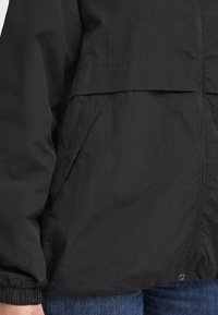 Svart zip-up jacka med elastiska ärmslut och sned sidficka, bärs över blå denimjeans.