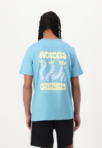 Gai zila kokvilnas T-krekls ar lielu dzeltenu un purpursarkanu grafiku mugurpusē, uz kuras ir uzraksts "ADIDAS ORIGINALS" un augu motīvi.