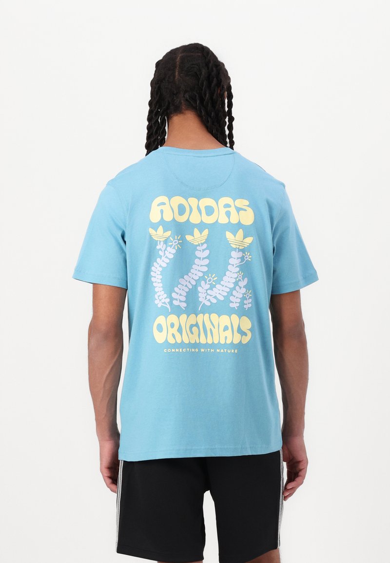 Gai zila kokvilnas T-krekls ar lielu dzeltenu un purpursarkanu grafiku mugurpusē, uz kuras ir uzraksts "ADIDAS ORIGINALS" un augu motīvi.