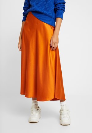 Vrouw draagt een felblauwe kabelgebreide trui, glanzende oranje midirok, witte sokken en stevige witte sneakers, staand tegen een effen achtergrond.