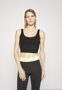Top corto nero in cotone con motivo testurizzato e spalline larghe, abbinato a leggings neri a vita alta con una fascia in giallo chiaro.