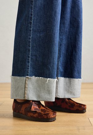 Scarpe marroni con motivo, dotate di una tomaia texture, indossate con jeans larghi in denim blu con un orlo sfilacciato di un grigio chiaro. Sfondo in legno.