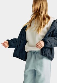 Granatowa kurtka puchowa o pikowanej fakturze, z zamkiem błyskawicznym i dwiema kieszeniami, na białym swetrze i jasnoniebieskich spodniach.