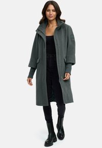 Manteau en polaire long et vert foncé avec un col montant, deux poches avant et une fermeture éclair. Associé avec un pantalon noir et des bottines.