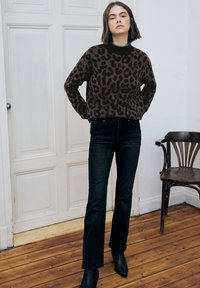 Brązowy sweter w panterkę z czarnym prążkowanym kołnierzem, do tego ciemne dzwoniaste dżinsy i czarne botki na drewnianej podłodze.