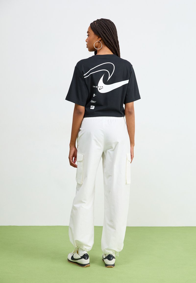Schwarzes Baumwoll-T-Shirt mit einem weißen, abstrakten Wellenmuster auf der Rückseite, kombiniert mit weißen Cargo-Hosen, die seitliche Taschen und elastische Bündchen haben.