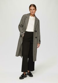 Zwart-wit houndstooth jas met een dubbelgebreid ontwerp, zijzakken en brede mouwen, gecombineerd met zwarte broek en glanzende instappers.