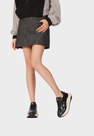 Donna con sneakers nere, gonna corta scura e giacca bicolore, in piedi con una gamba incrociata davanti all'altra.