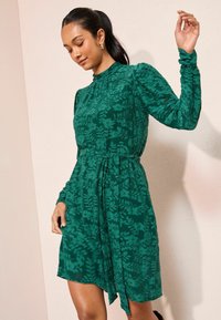 Zelené šaty vyrobené z texturované látky se vzorem kapradiny, s vysokým výstřihem, dlouhými rukávy a detailem zavazování v pase s asymetrickým lemem.