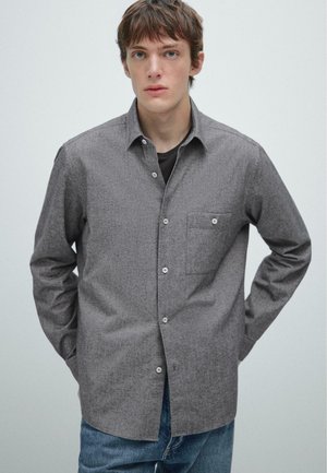 Camicia grigia con bottoni e tessuto testurizzato, taschino sul petto, e colletto classico, abbinata a jeans blu.