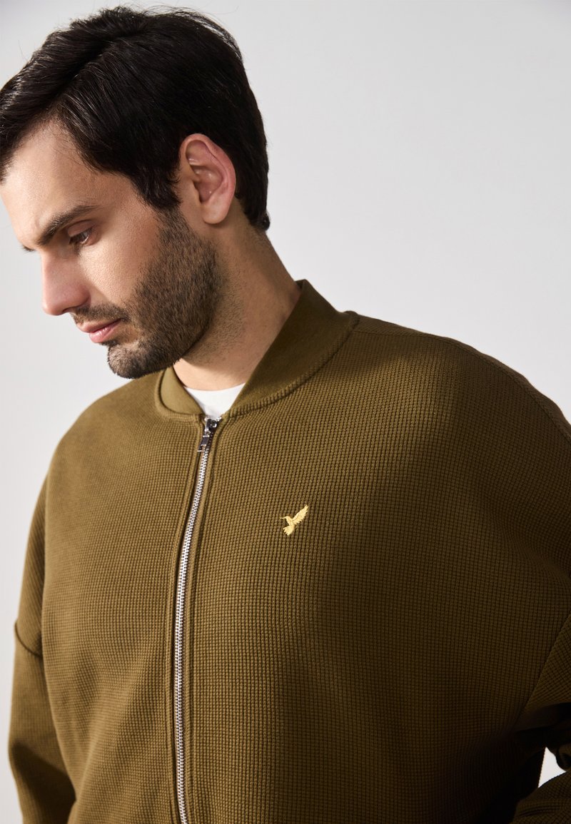 Olivgrüne Zip-Jacke mit strukturiertem Rippmaterial, goldfarbenem Logo-Detail auf der Brust und Stehkragen. Glattes Material und lässiger Schnitt.