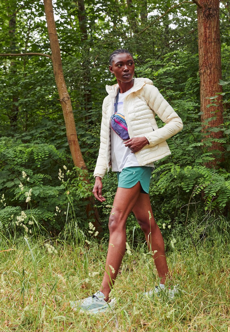 Chaqueta de plumas beige claro, pantalones cortos verdes, camiseta blanca, bolso cruzado pequeño multicolor. Modelo caminando en un entorno forestal con hierba.