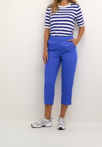 Blaue, cropped Hosen mit glatter Textur, kombiniert mit einem kurzärmligen Oberteil in Weiß und Blau mit Streifen. Getragen mit grauen und weißen Sneakern.