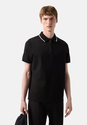 Jonge man met kort haar draagt een zwart polo met korte mouwen en witte rand, houdt een zwarte pet vast, staat tegen een effen lichte achtergrond.