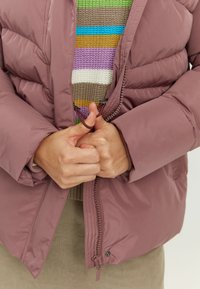 Puffer jacket in muted pink, featuring a zip closure and quilted texture. Layered underneath is a multicolored striped knit sweater.
