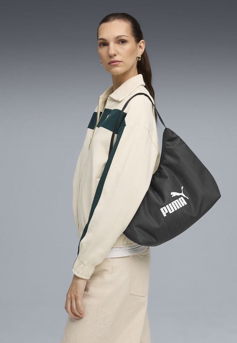 Borsa a tracolla grigia con texture e strap nera, con un grande logo Puma bianco in evidenza. Il modello indossa una giacca crema con strisce verdi e una gonna beige.