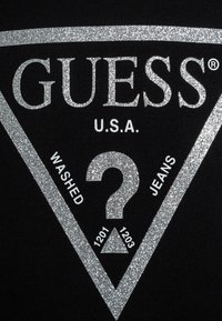 Tela negra con un logo y texto plateados brillantes dentro de un diseño triangular. El texto incluye "GUESS", "EE.UU.", "LAVADO" y "JEANS."
