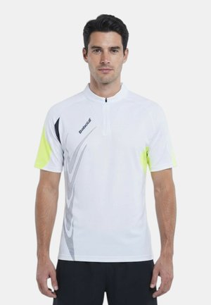 Uomo che indossa una maglia sportiva bianca a maniche corte con mezza cerniera, accenti neri e gialli e pantaloncini neri, in piedi rivolto in avanti.