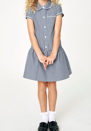 Enfant portant une robe à carreaux noirs et blancs, des chaussettes blanches à volants et des chaussures noires, debout les mains jointes devant.