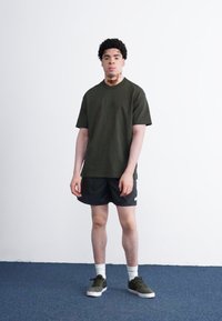 T-shirt i mørkegrøn bomuld, rund hals, korte ærmer. Kombineret med sorte shorts, hvide ankelstrømper og mørkegrønne sneakers. Står på blå tæppe.