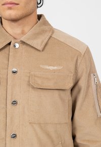 Jasno brązowa kurtka z kordurowego materiału, z kołnierzem, srebrnymi guzikami, kieszenią na piersi oraz haftowanym logo z detalami skrzydeł.