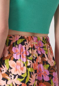 Zeleni rebrasti crop top u kombinaciji s živopisnim cvjetnim hlačama u ružičastoj, narančastoj, ljubičastoj i crnoj boji, s naglašenim strukom i suptilnim logotipom.