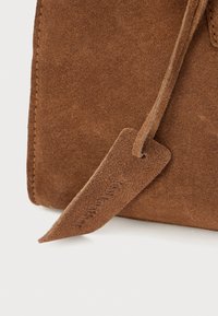 Coin d'un sac en suédine marron avec une finition texturée et une longue bandoulière plate. Détails de l'étiquette visibles, avec texte imprimé et bords doux.