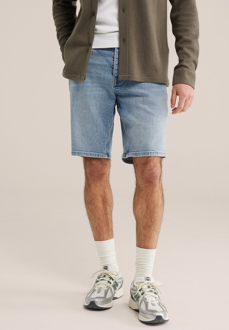 Mees, kes kannab helesiniseid teksapükse, valgeid sokke, halli ja rohelistes toonides tossusid, valget särk ja oliivirohelist pika varrukaga nööbitavat särki.