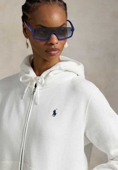 Femme portant un sweat à capuche blanc zippé avec un logo joueur de polo bleu marine, de grandes lunettes de soleil bleu foncé et des boucles d'oreilles pendantes en coquillage, regardant vers la gauche.