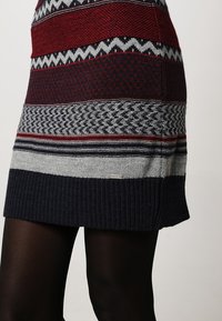 Jupe en tricot avec des motifs horizontaux bordeaux, gris et bleu marine portée avec des collants noirs sur un fond neutre.