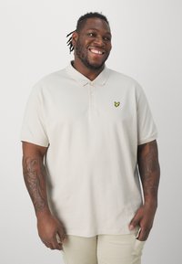 Beige poloko s límečkem, krátkými rukávy a žlutým vyšívaným logem. Materiál vypadá měkce; hladká textura s uvolněným střihem.