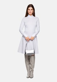 Weißes langärmeliges Kleid mit plissiertem Rock, kombiniert mit einer silbernen Handtasche. Das Model trägt graue Stiefel bis zum Knie. Glatte Textur, tailliertes Oberteil.