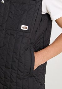 Veste matelassée noire North Face portée sur une chemise blanche, main insérée dans la poche latérale droite.